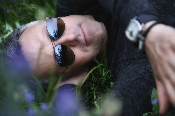 ハンサムな中年の男性の肖像画 青の花の間でフィールドに横たわっていた 自然の屋外でのプロファイルでは無気力な穏やかな男 緑の草の中に閉じられた男性の顔 — ストック写真