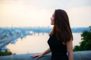 Ukrayna 'nın başkenti Kyiv' deki Dniper nehrinin kıyısında akşam poz veren siyah şık elbiseli manken. Genç, güzel esmer, beyaz kadın yaz sokaklarında yürüyor. Güzel kız, şehir portresi..