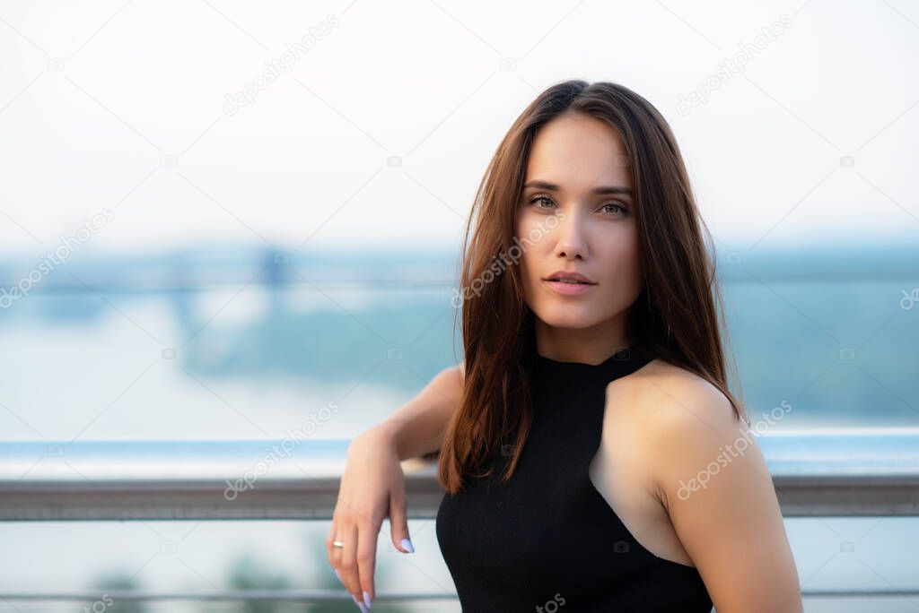 Fashion model wearing black elegant dress posing outdoor at the bank of Dniper river in Kyiv, Ukraine. Young beautiful brunette caucasian woman walking summer streets. Beautiful girl, urban portrait.