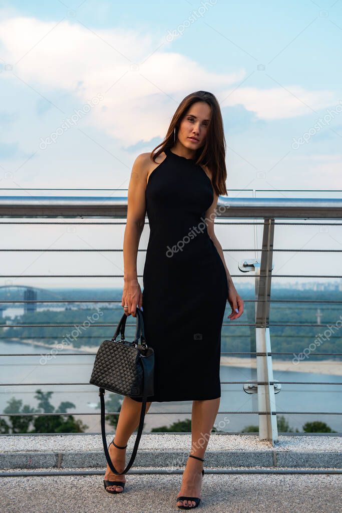 Fashion model wearing black elegant dress posing outdoor at the bank of Dniper river in Kyiv, Ukraine. Young beautiful brunette caucasian woman walking summer streets. Beautiful girl, urban portrait.