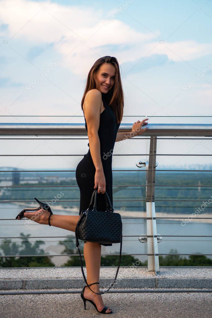 Fashion model wearing black elegant dress posing outdoor at river Dniper bank in Kyiv, Ukraine. Young beautiful brunette caucasian woman walking summer streets. Beautiful girl, urban portrait.