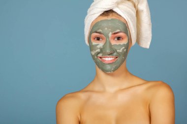 Yüzüne kil maskesi takan güzel, neşeli bir kız. Mavi arka planda genç kadın ve güzellik tedavileri. Şımartma, gençlik, yaşlanma karşıtı, vücut bakımı spa teması. Kozmetik maskeli kadın yüzü