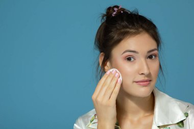 Neşeli, çekici genç kız yüzünü pamuk diskle temizliyor, mavi arka planda izole edilmiş. Sabah ameliyatları sırasında güzel bir kadının portresi.