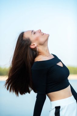 Çekici, gülümseyen esmer kız dışarıda göz kırpıyor. Genç kadın, yaz portresi. Doğada nehir, sahil ve mavi temiz gökyüzü ile poz veren kadın model..