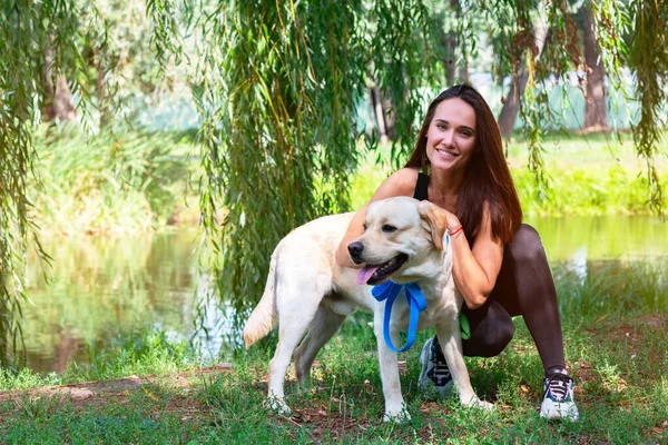 陽気なかなり若いです女性座っていると抱擁彼女の犬で川の銀行で夏の公園 — ストック写真