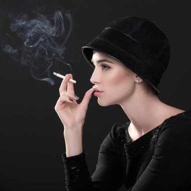 Şapkalı sigara içen genç bir moda kadını ve esmer arka planda siyah süveterler. Klasik kadın portresi, tasarım. Resim tonlandırılmış.