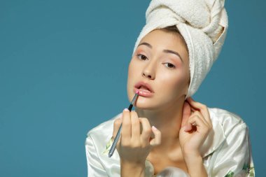 Genç ve çekici bir kız mavi arka planda dudaklarına ruj sürüyor, güzellik tedavi sabahı. 