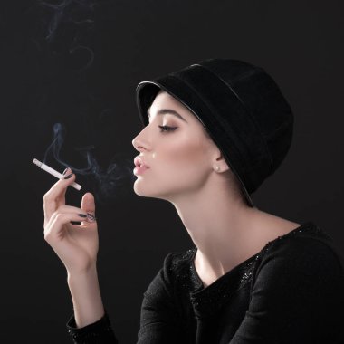 Şapkalı sigara içen genç bir moda kadını ve esmer arka planda siyah süveterler. Klasik kadın portresi, tasarım. Resim tonlandırılmış.