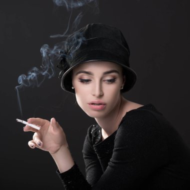Şapkalı sigara içen genç bir moda kadını ve esmer arka planda siyah süveterler. Klasik kadın portresi, tasarım. Resim tonlandırılmış.
