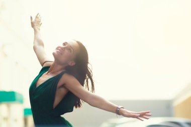 Dışarıda yaz şehrine poz veren sevimli, neşeli genç bir kadın. Kendine güveni olan, zümrüt renkli tulum giymiş, uzun kahverengi saçlı, hayat dansı yapan güzel bir kız. Kentsel yaşam tarzı, rüzgarlı hava, görüntü tonlu