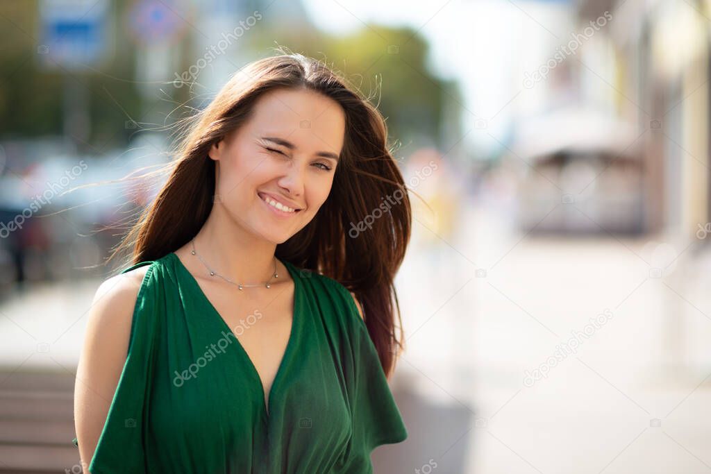 Young pretty likable cheerful woman posing summer city outdoor. Beautiful self-confident girl dressed in emerald-colored jumpsuit with long brown hair walking street enjoing her life, urban lifestyle