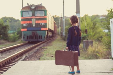 At kuyruklu güzel bir kız tren istasyonunda bekliyor. Koyu mavi elbiseli, çiçekli, bluzlu, elinde büyük bir bavul var. Genç gezgin, eski moda. Dışarıdaki şirin çocuk portresi.