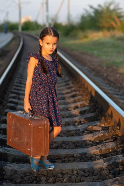 Beautiful charming little girl with pigtails dressed in dark blue dress with flowers stands at railroad with big vintage luggage. Fashion, retro stylization.