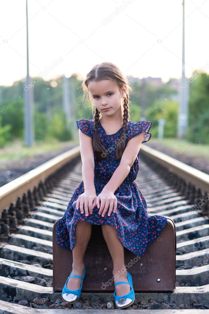 Beautiful charming little girl with pigtails dressed in dark blue dress with flowers and blouse sitting in big vintage luggage at railroad. Fashion, retro stylization. 