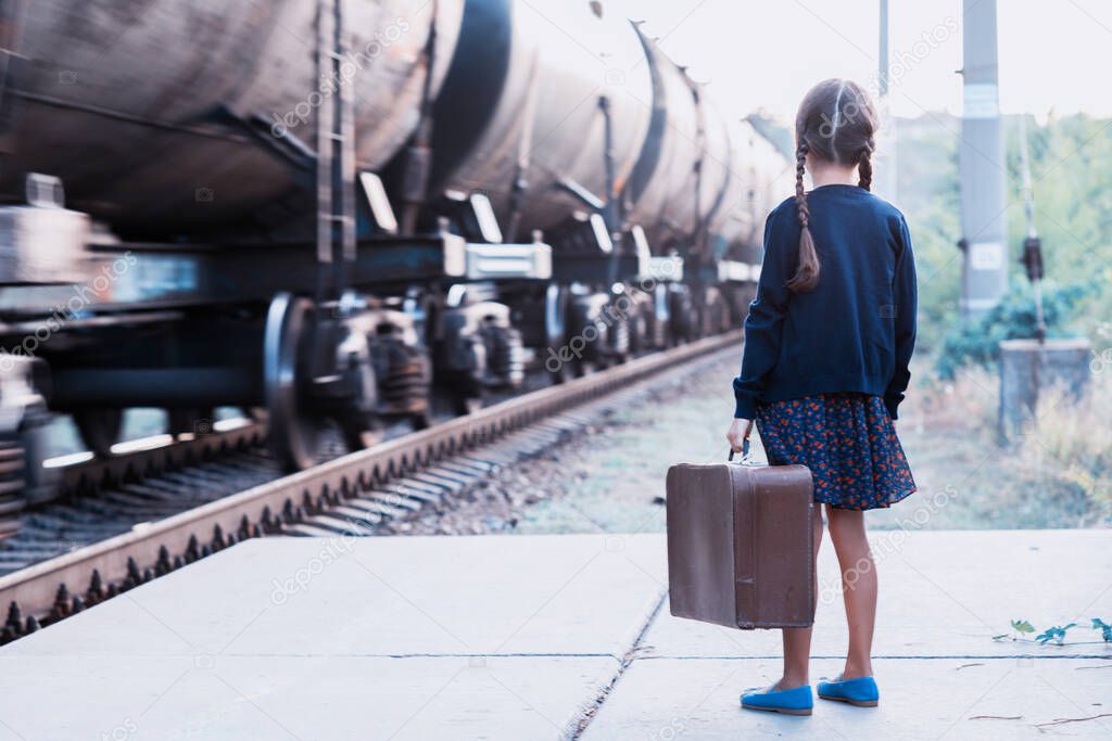 Beautiful charming little girl with pigtails waiting for train at station dressed dark blue dress with flowers and blouse holding big vintage luggage. Young traveler, retro stylization. Cute kid outdoor portrait
