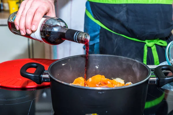 Macho mão segurando garrafa e derramando vinho tinto em caçarola pan com fritar cenouras e cebolas no fogão elétrico moderno — Fotografia de Stock