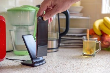 Akıllı telefon, mutfak masasının üstündeki şarj masasında şarj oluyor. Erkek el telefonu şarja yerleştiriyor