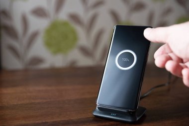 Akıllı telefon ahşap masadaki şarj masasında şarj oluyor. Erkek el telefonu şarja yerleştiriyor