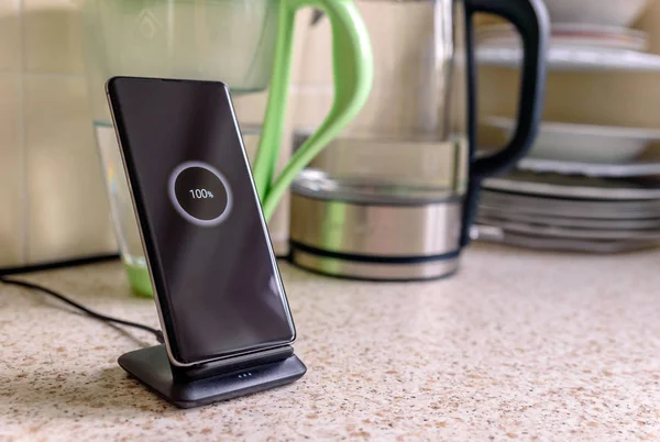 Smartphone carga inalámbrica en el soporte de carga con el icono del 100 por ciento en la pantalla en la mesa de la cocina — Foto de Stock