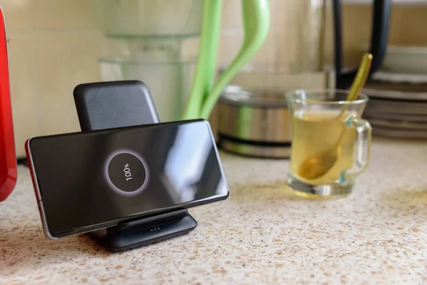 smartphone wireless charging on charging stand with 100 percent icon on screen on kitchen tabletop