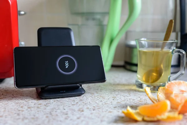 Smartphone carga inalámbrica en el soporte de carga con el icono del 100 por ciento en la pantalla en la mesa de la cocina junto a la taza de vidrio del té en la mesa de la cocina — Foto de Stock