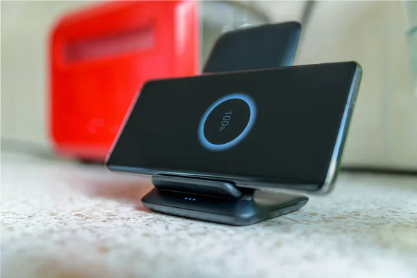 Smartphone carga inalámbrica en el soporte de carga con el icono del 100 por ciento en la pantalla en la mesa de la cocina junto a la tostadora — Foto de Stock