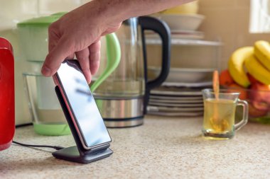 Akıllı telefon, mutfak masasının üstündeki şarj masasında şarj oluyor. Erkek el telefonu şarja yerleştiriyor