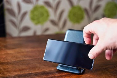 Akıllı telefon ahşap masadaki şarj masasında şarj oluyor. Erkek el telefonu şarja yerleştiriyor