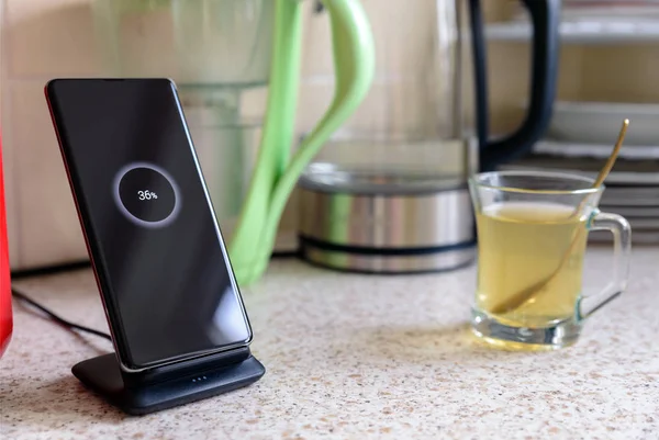 smartphone wireless charging on charging stand with 36 percent icon on screen next to tea glass cup on kitchen tabletop
