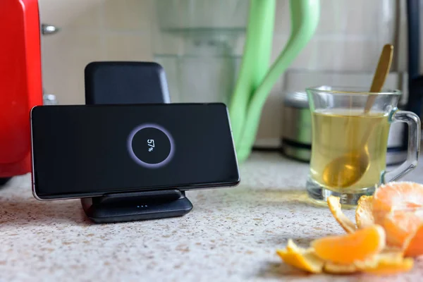 Smartphone carga inalámbrica en el soporte de carga con 57 por ciento icono en la pantalla en la mesa de la cocina junto a la taza de vidrio de té en la mesa de la cocina — Foto de Stock