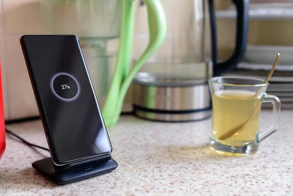 Carga inalámbrica de teléfono inteligente en el soporte de carga con el icono del 27 por ciento en la pantalla junto a la taza de vidrio de té en la mesa de la cocina — Foto de Stock
