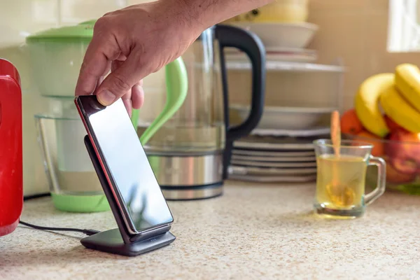 smartphone wireless charging on charging stand on kitchen tabletop. male hand placing phone to charging