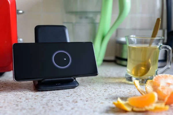 Smartphone carga inalámbrica en soporte de carga junto a la taza de vidrio de té en la mesa de la cocina — Foto de Stock