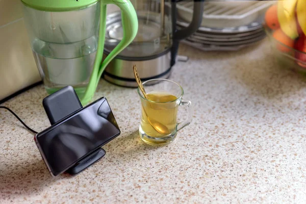 smartphone wireless charging on charging stand next to tea glass cup on kitchen tabletop