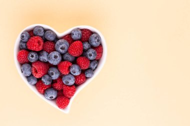 Yaban mersini ve ahududu, kalp şeklinde bir tabakta meyve porselen bir çanakta pastel arka planda