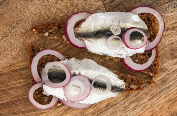 Sprat op een stuk zwart brood. Vis met brood en rode ui op een snijtafel. — Stockfoto