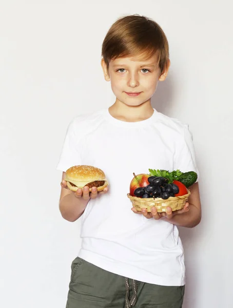 Een blonde jongen kiest tussen een hamburger, verse groenten en fruit. — Stockfoto