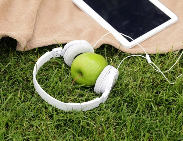 Headphones, electronic tablet and green apple for picnic in park on green grass