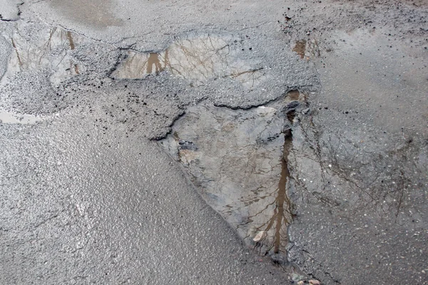 Tehlikeli yol yatağı yok edildi. Büyük bir çukur ve suyla dolu kötü asfalt yol. Sudaki ayna yansıma ağaçları.