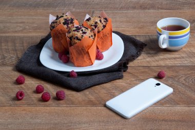 Ahududulu üç lezzetli kek beyaz bir tabakta yatıyor. Sabah kahvaltısı. İletişim için bir fincan çay ve akıllı telefon..