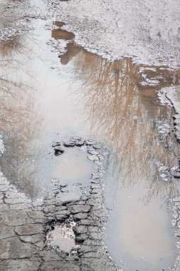 Kötü asfalt yol. Suyla dolu büyük bir çukur. Tehlikeli yol yatağı yok edildi. Sudaki ayna yansıma ağaçları.