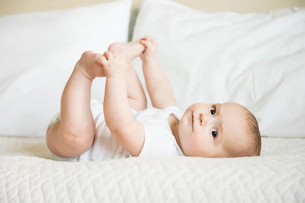 Happy baby boy in white onesie holds feet — Stock Photo, Image