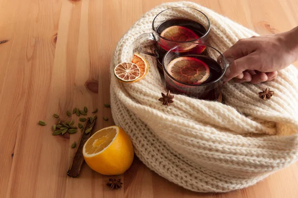 Seasonal and holidays concept . Christmas mulled wine with beautiful orange slices inside the glass ,covered with warm white scarf .Warm and cozy atmosphere.Men holding on of the cups with right hand. — Stock Photo, Image