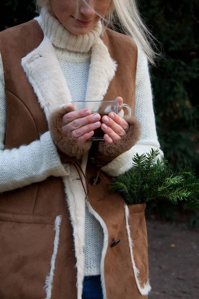 Το κορίτσι κάτω από το ζεστό, καφέ παλτό με μεγάλη τσέπη έχει κλαδιά από έλατο στο εσωτερικό του. Όλοι μαζί να κάνει την εντύπωση ότι είναι μετά από harving έλατο στο δάσος για στεφάνι με Chriistmas. Και μετά από αυτό αυτή ζεσταίνει τα χέρια της με ζεστό ρόφημα — Φωτογραφία Αρχείου