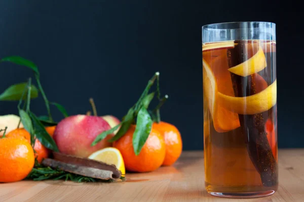Glass with Seasonal warming drink.Cinnamon,lemon zest and some fruits inside.On the wooden table.With mandarine,apple and orange on the back. — Stock Photo, Image