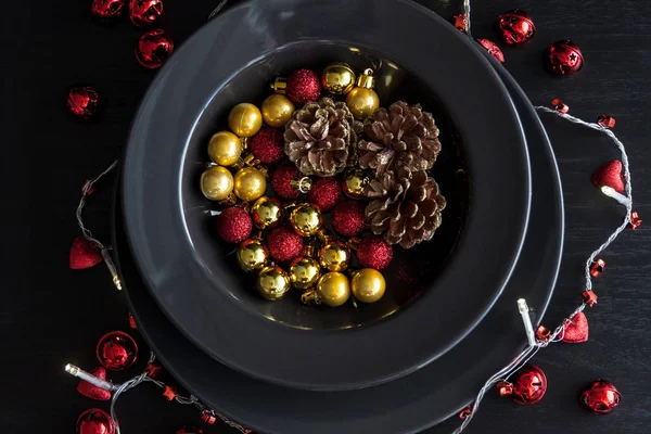 Weihnachtsschmuck in der schwarzen Platte auf dunklem Hintergrund. Rot, goldene Kugeln und Zapfen innen und Weihnachtsbeleuchtung ringsum. — Stockfoto