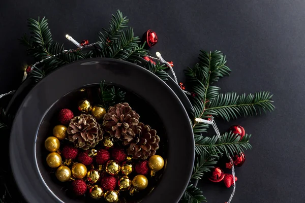 Christmas Decorations in the black plate on the dark background with some spruce twigs.Red, gold balls and cones inside and christmas light around . — стоковое фото