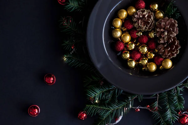 Christmas Decorations in the black plate on the dark background with some spruce twigs.Red,gold balls and cones inside and christmas light around. — Stock Photo, Image