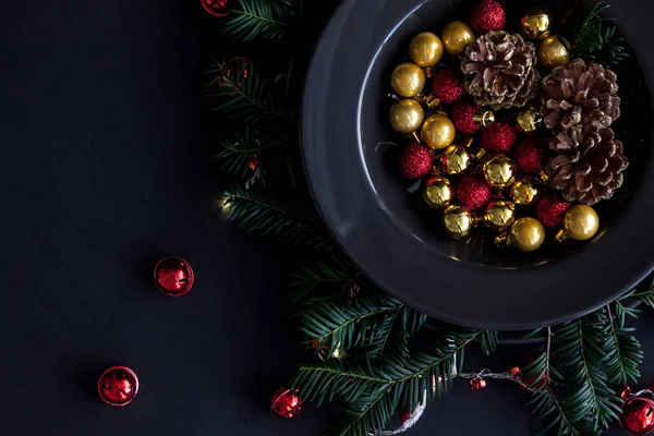 Christmas Decorations in the black plate on the dark background with some spruce twigs.Red, gold balls and cones inside and christmas light around . — стоковое фото