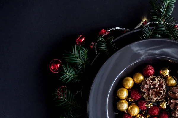 Christmas Decorations in the black plate on the dark background with some spruce twigs.Red, gold balls and cones inside and christmas light around . — стоковое фото